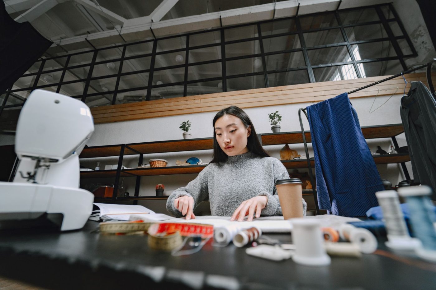 A focused fashion designer works in a studio, surrounded by sewing tools, cloth, and a coffee cup.
